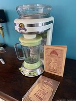Margaritaville DM 1000-33 Frozen Concoction Maker with instruction manual and recipe booklet on wooden surface against blue wall.