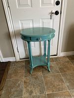 Small teal round occasional table with drawer, four legs, bottom shelf, and glass drawer knob shown against white door and tiled floor.