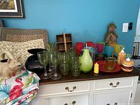 Photo of assorted plastic glasses, novelty drink containers, colorful table linens, and part of a black CD player/radio on a dark wood sideboard against a blue wall.