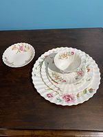Stacked 6 piece Clovelly place setting showing the cup, saucer, bread, salad, dinner plates and bowl with floral pattern and scalloped edges on a wooden surface.