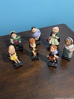Seven Royal Doulton Dickens character ceramic figurines displayed upright on a wooden surface showing front details and facial expressions.