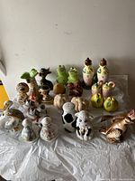Photo showing full lot of assorted salt and pepper shakers arranged on a surface with a yellow tool cabinet in the background. The photo highlights the size, quantity, and variety of the ceramic figures including frogs, birds, and fruits.