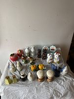Photo of 12 sets of salt and pepper shakers arranged on a white cloth with various animal and object shapes visible.
