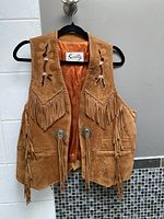 Front view of the tan suede leather vest showing beadwork near the shoulders, fringe detailing, front pockets, conchos with leather ties, and orange lining inside.