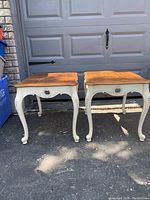 Pair of Ethan Allen side/end tables with wooden tops and carved white legs shown outdoors on concrete in front of a garage door.