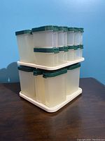 Photo of two-tier Tupperware spice carousel set on a dark wooden surface against a light blue wall, showing translucent containers with green lids arranged on white rotating trays.