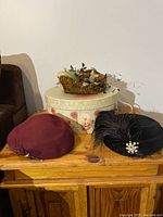 Three vintage hats displayed on a wooden surface with a floral hat box behind them. Hats include a burgundy fur felt hat, a black feathered hat with pearl embellishment, and a multi-colored feathered pillbox hat.