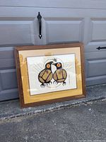 Photo of a framed numbered print showing two stylized birds with vibrant colors within a wooden frame placed against a garage door.