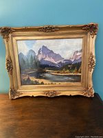 Framed oil painting of mountain landscape with river and trees against blue wall, on wooden surface.
