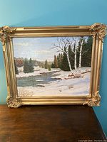 Framed oil painting showing a winter scene with snow, river, birch and pine trees in outdoor natural setting.