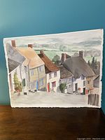 Watercolour painting leaning against a teal wall, showing village houses and distant hills.