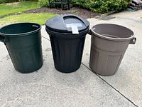 Set of three plastic trash cans in green, black with lid, and brown with handles placed on concrete outdoor driveway area.