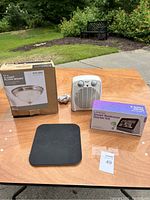 Overview of the lot showing all items on a wooden table outdoors: Comfort Zone heater, Lenovo Smart Bedroom Starter Kit box, Hampton Bay ceiling light box, and black electronic device with 3 circular touch zones.