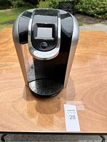 Front view of black Keurig coffee maker with digital screen and water reservoir on the side, placed on wooden surface outdoors.