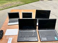 Photo showing 5 assorted laptops on a wooden table outdoors: Dell Inspiron, HP, Samsung, and two smaller laptops.