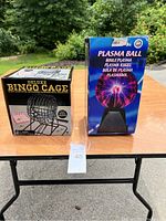 Photo showing plasma ball box and deluxe bingo cage box side by side on a black table outdoors with greenery background.