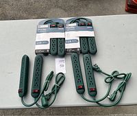 Photo showing six green power strips on white table, including two new GE six-outlet power strips with packaging and four used green power strips with multiple outlets and cords.