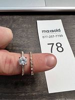 Photo showing two rings held on finger against auction number card. One ring has a large central clear stone; the other is a band with smaller clear stones.