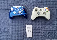 Photo shows two Xbox wireless controllers placed on a dark quilted surface, one translucent blue and one white, along with a MaxSold auction lot tag.