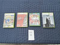 Four Xbox game cases laid out on a blue quilted fabric surface. Visible titles are Sonic Sega Superstars Tennis, Project Gotham Racing 3, The Sims 4, and Batman Arkham Asylum.