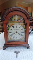 Full front view of antique wooden mantle clock with arched top, hand-painted metal face, Roman numerals, and key in front