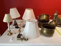 Overview of all brass lamps with shades, brass pots, and coffee pot on table against red wall.
