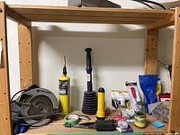 Photo showing circular saw, plunger, yellow flashlight, tape, and small hardware items on wooden shelves