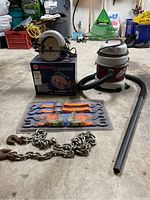 Lot items on garage floor showing circular saw on its box, shop vac with hose and wand, tie down straps in packaging, and heavy chain.
