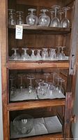 Multiple rows of clear glass stemware including wine glasses and goblets displayed in wooden cabinet with clear glass bowl also visible