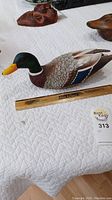 Hand carved wooden duck decoy on white quilted surface with ruler showing length around 12 inches.