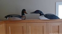Two hand carved wood waterfowl figurines, a duck and a loon, side by side on a wooden cabinet, showing overall size and shape