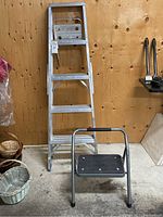 Photo showing 5-foot aluminum step ladder against wooden wall with metal step stool in front, some baskets to the left and tool storage on right.