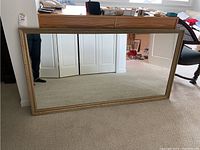 Front view of large rectangular mirror leaning against a wall showing gold frame and mirror surface with some marks