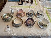 Overview of seven tea cup and saucer sets and one small creamer pitcher arranged on table.