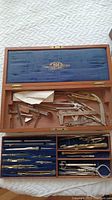 Top view of open wooden case showing various precision measurement tools including compasses and calipers arranged on blue velvet lining.
