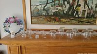 Wide photo showing all glass and crystal stemware, hand painted platter on left with floral design, multiple clear glasses and mugs arranged on wooden mantelpiece