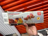Hand holding original packaging box of battery operated super peeler showing the product image, name, and 'As Seen On TV' logo.