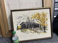 Photo showing the embroidered cloth painting of a covered bridge with yellow trees and birds, placed on the ground next to other frames for size reference.