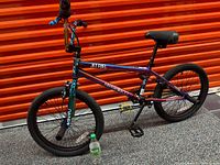 Side view of BMX bike with gradient multicolor frame, black tires, black seat, blue handlebar grips, and yellow pedals featuring a Sprite bottle on the floor for scale.