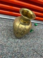 Front angle view of the brass flower pot standing on carpet, showing the hammered texture and hanging chains.