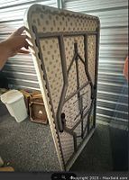 Side view of the folding table held upright showing underside with black metal legs folded against plastic top