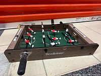Side angle view of one mini foosball table showing woodgrain frame, green field, plastic players in red and white, with black handles and Brookstone branding.