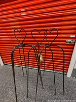 Four black metal garden planter stands with heart-shaped tops, standing together against an orange background.