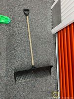 Photo of heavy duty snow shovel with wooden handle and black plastic scoop laid on carpeted floor.
