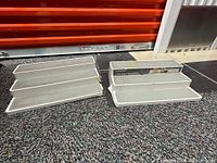 Photo showing two separate groups of white plastic tiered kitchen shelf racks placed on a carpeted floor with an orange shutter in the background.