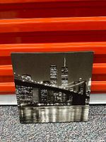 Front view of monochrome painting of city bridge at night with skyline and water reflection against an orange background.