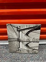 Square black and white canvas painting showing an inside view of a suspension bridge with cables and beams.