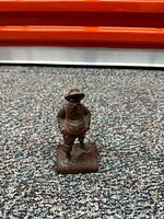 Photo showing a small bronze soldier or worker figurine standing on a carpeted floor with a bright orange object in the background.