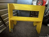 Front view of yellow wooden bench showing rectangular open backrest and seat surface.