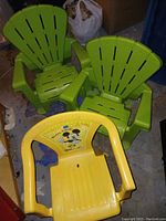 Two green plastic Adirondack style toddlers chairs and one yellow toddlers chair with Mickey Mouse graphic together on a floor.